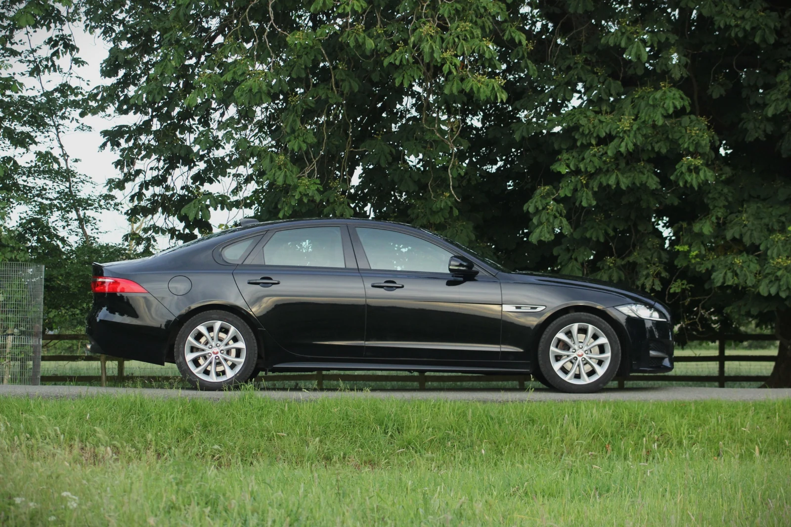 The full side profile of a black Jaguar on a drive beside grass and trees.